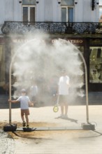 A mobile spray arch sprays a fine mist of water on a sunny square. Passers-by, including a child