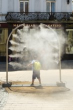 A mobile spray arch sprays a fine mist of water in a sunny square. A child uses the refreshment in