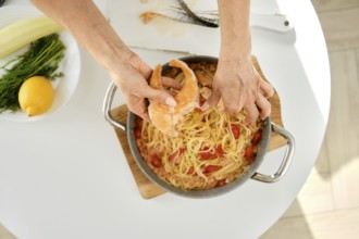 A person is adding salmon to a pot of pasta with tomatoes and herbs on a sunny kitchen countertop.