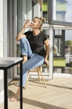 A middle-aged woman sits comfortably on a wooden chair in a sunny balcony, sipping from a glass.