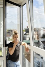 A woman leans against a balcony railing, sipping a drink and looking out at the vibrant cityscape.