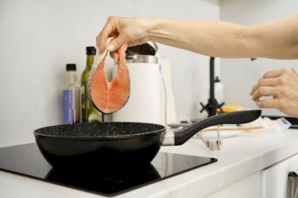 A hand is lifting a fresh salmon fillet above a black frying pan on a stovetop. The kitchen appears