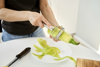 A person is peeling a zucchini in a bright kitchen. Fresh zucchini is held in one hand while a