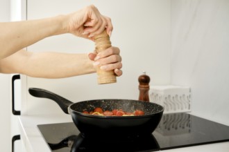 A hand is grinding pepper over a skillet filled with fresh tomatoes. The setting is a modern