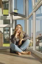 A middle-aged woman sits comfortably on a wooden floor of balcony in a bright modern apartment. She
