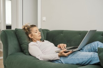 A woman with a relaxed demeanor is laying on a green couch in a bright living room, focused on her