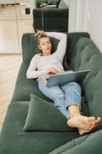 Middle-aged woman is lounging on a green couch, scrolling on her laptop in online shop choosing