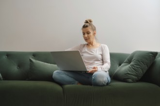 A woman sits cross-legged on a soft green couch, focused on her laptop. Natural light fills the