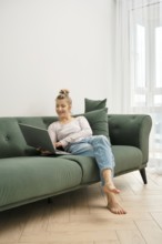 A woman enjoys her time on a green couch, focused on her laptop. Sunlight fills the bright living