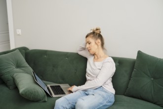 Middle-aged woman is seated on a green couch in a well-lit living room, working intently on her