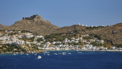 Coastal town on a blue sea with hill and castle in the background, lined with sailing boats,