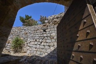 View through open Tor tor to stone wall crowned by flowers, blue sky visible, Open entrance gate,