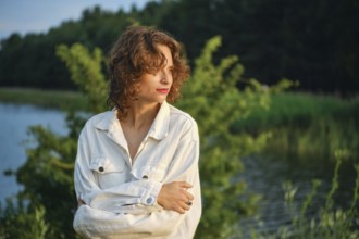 On a hot day, a woman stands near the calm lake, basking in the sunlight. Surrounded by lush