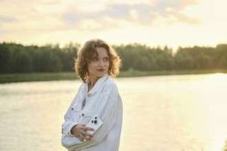 Cute woman with curly hair stands by the lake at sunset. She look back while holding smartphone in