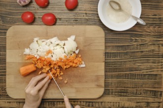 A person cutting diced carrots and onions on a wooden cutting board. Fresh tomatoes and garlic can