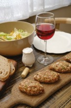 A table is set with a wooden board of homemade patties, a bowl of fresh salad, a glass of red wine,