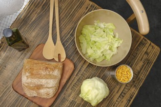 A rustic wooden table displays a loaf of bread, a bowl of chopped lettuce, and a cup of corn.