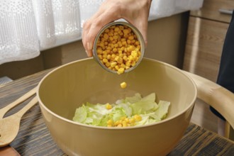 A hand pours canned corn into a bowl filled with fresh green lettuce. The warm kitchen features