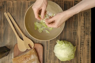 Hands tear crisp lettuce leaves into a bowl, surrounded by wooden utensils and fresh ingredients.