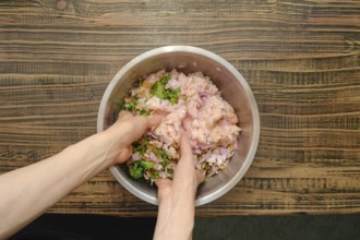 Hands are mixing raw minced meat, chopped onions, herbs, and spices in a stainless steel bowl on a