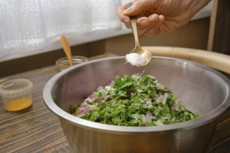 A hand adds salt to a bowl filled with minced meat, chopped onions and fresh herbs. The kitchen is