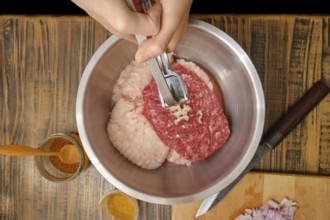 A hands use press to add minced garlic to a mixture of ground meats in a stainless-steel bowl.