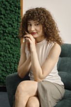 Cute woman with curly hair sits gracefully on a green armchair, resting her chin on her hands. She