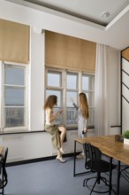 Two women engage in a conversation while standing by large windows in a modern office setting. The
