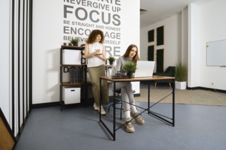 Two women working in bright office. One takes notes while the other types on a laptop, showcasing