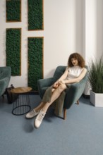 A young woman sits comfortably in a contemporary chair, dressed in a casual outfit. The room