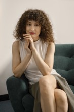 A woman with curly hair smiles while seated on a green couch. She wears a light top and a skirt,