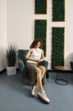 A fashionable woman is seated in a chair within a contemporary room adorned with lush green wall