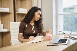 A woman sits at a wooden desk in a stylish home office, writing in a notebook while dropping coins