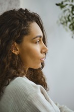 In a cozy indoor space, a woman with long, curly hair sits in profile, lost in thought. Soft