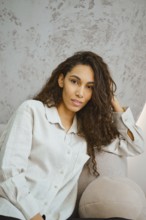 Charming woman with curly hair sits comfortably on a plush sofa in a contemporary indoor space.