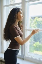 A woman admires the greenery outside while standing by a window. Her curly hair cascades down her