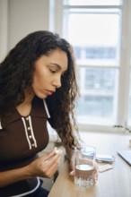 A young woman with curly hair sits at a table concentrating on a pill in her hand and a glass of