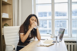 A focused woman sips from a glass of water at her desk during a work session in a well-lit office.