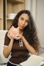 A portrait of a young woman with curly hair holding a cute pink piggy bank. She appears focused and