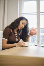 A woman with long curly hair sits at a wooden desk, calculating vacation expenses in a notebook. In