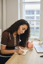 A woman with curly hair writing in her notebook at a wooden desk. She organizes coins beside a pink