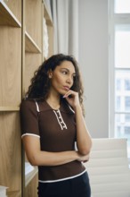 A woman with curly hair stands pensively in a well-decorated office. She rests against wooden