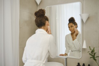 A woman in a white robe is in a sleek bathroom, gently washes makeup off her face. She focuses on