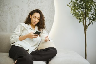 A young woman sits comfortably on a couch in a modern living room, focused on her smartphone while
