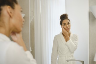 A woman in a white bathrobe stands in front of a mirror, applying skincare to her face. The bright,