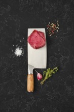 Fresh meat sits atop a large knife on a dark countertop, surrounded by salt, pepper, herbs, and