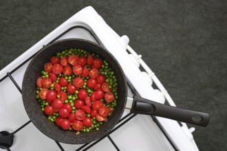 A skillet on a gas stove with red cherry tomatoes and green peas, cooking together in garlic butter