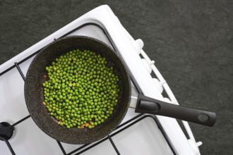 Fresh green peas are being cooked in a black pot on a white stovetop. Steam rises gently,
