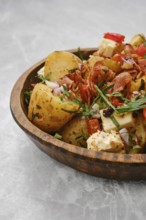 A bowl filled with cooked potatoes mixed with fresh vegetables, herbs, bacon and cheese. The dish