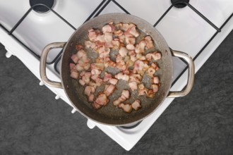 Bacon strips are sizzling and browning in a stainless steel frying pan placed on a stovetop. The